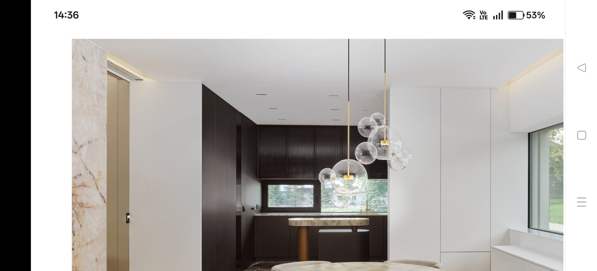 Nowoczesne wykończenie wnętrza: ciemna zabudowa kuchenna z oknem, marmurowa ściana, designerska lampa wisząca z szklanych kul i jasny stół. Minimalistyczny styl.