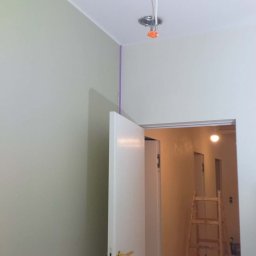 STRATUM GROUP - Wnętrze w trakcie prac wykończeniowych: świeżo pomalowane ściany w odcieniach szarości, widoczne przewody elektryczne wystające z sufitu, otwarta biała stolarka drzwiowa.