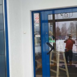 STRATUM GROUP - Biała elewacja budynku z niebieskimi drzwiami wejściowymi. W odbiciu szyby widoczny pracownik na drabinie. Podłoga wyłożona brązowymi płytkami.