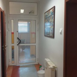 STRATUM GROUP - Korytarz po odświeżeniu: białe ściany, obraz na ścianie, białe drzwi z żaluzjami, grzejnik. Podłoga z szarym pasem na środku i pomarańczowymi brzegami.