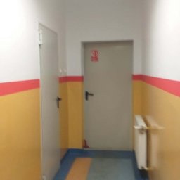 STRATUM GROUP - Korytarz po odświeżeniu: żółte ściany z czerwonym pasem, szare drzwi i podłoga w odcieniach niebieskiego i pomarańczowego. Widoczny grzejnik i oświetlenie sufitowe.