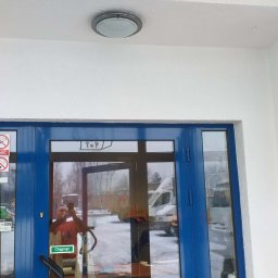 STRATUM GROUP - Błękitne drzwi wejściowe po malowaniu, widoczna drabina i odbicie osoby robiącej zdjęcie. Świeżo pomalowana elewacja na biało, okrągła lampa sufitowa.