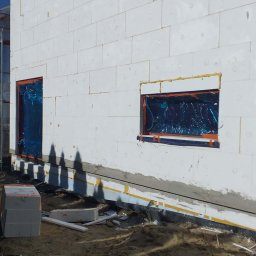 STRATUM GROUP - Ściana budynku w trakcie ocieplania, widoczne białe bloki styropianowe, zabezpieczone okna folią, przygotowane podłoże. Etap prac ociepleniowych.