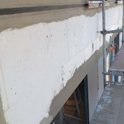 STRATUM GROUP - Ocieplona ściana budynku w trakcie prac wykończeniowych, widoczne rusztowania i zabezpieczone okna folią. Szara warstwa szpachli na białym styropianie.