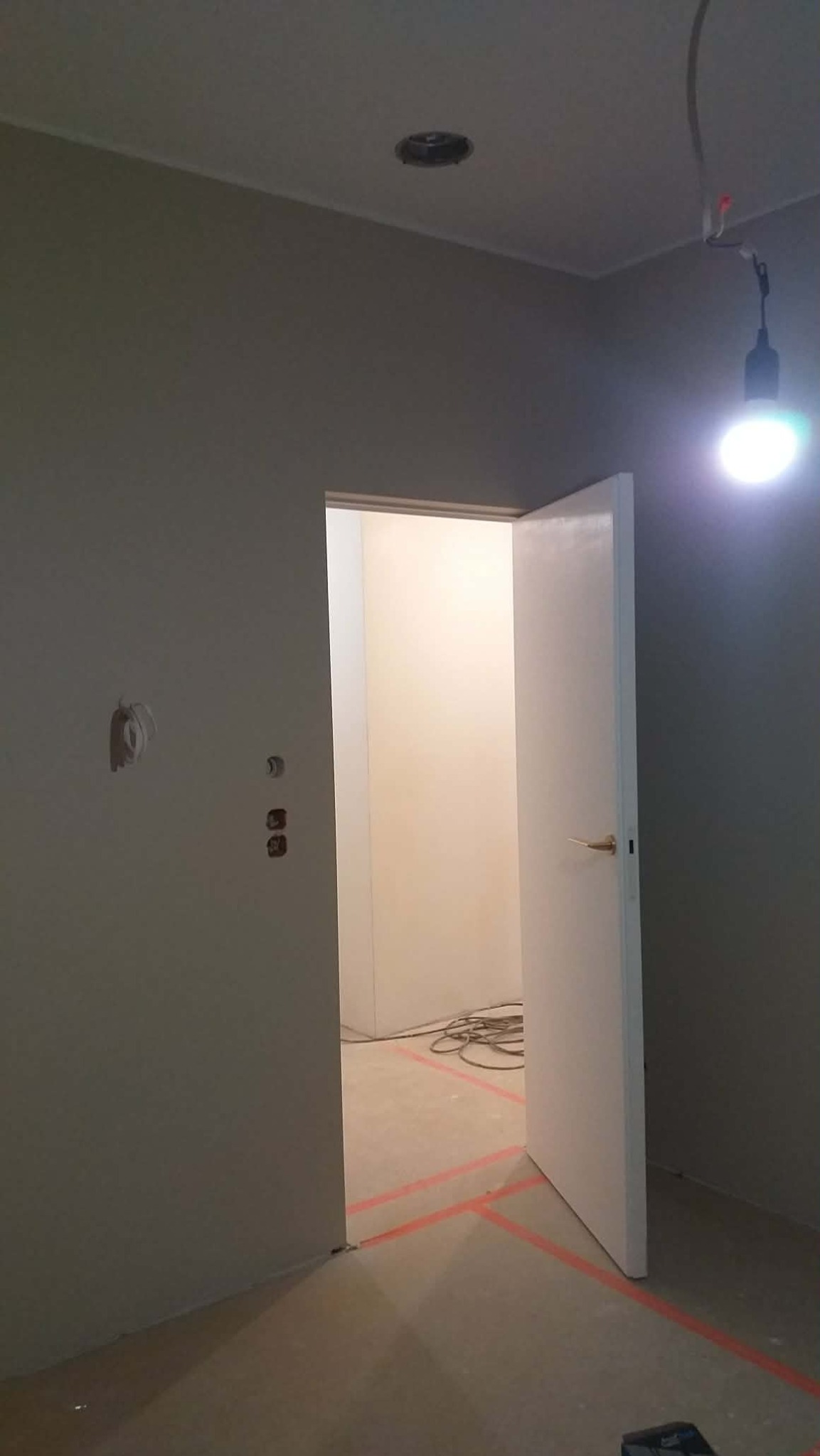 Wnętrze w trakcie prac wykończeniowych: szare ściany, białe drzwi, widoczne instalacje elektryczne i oznaczenia na podłodze. Otwór w suficie i prowizoryczne oświetlenie.