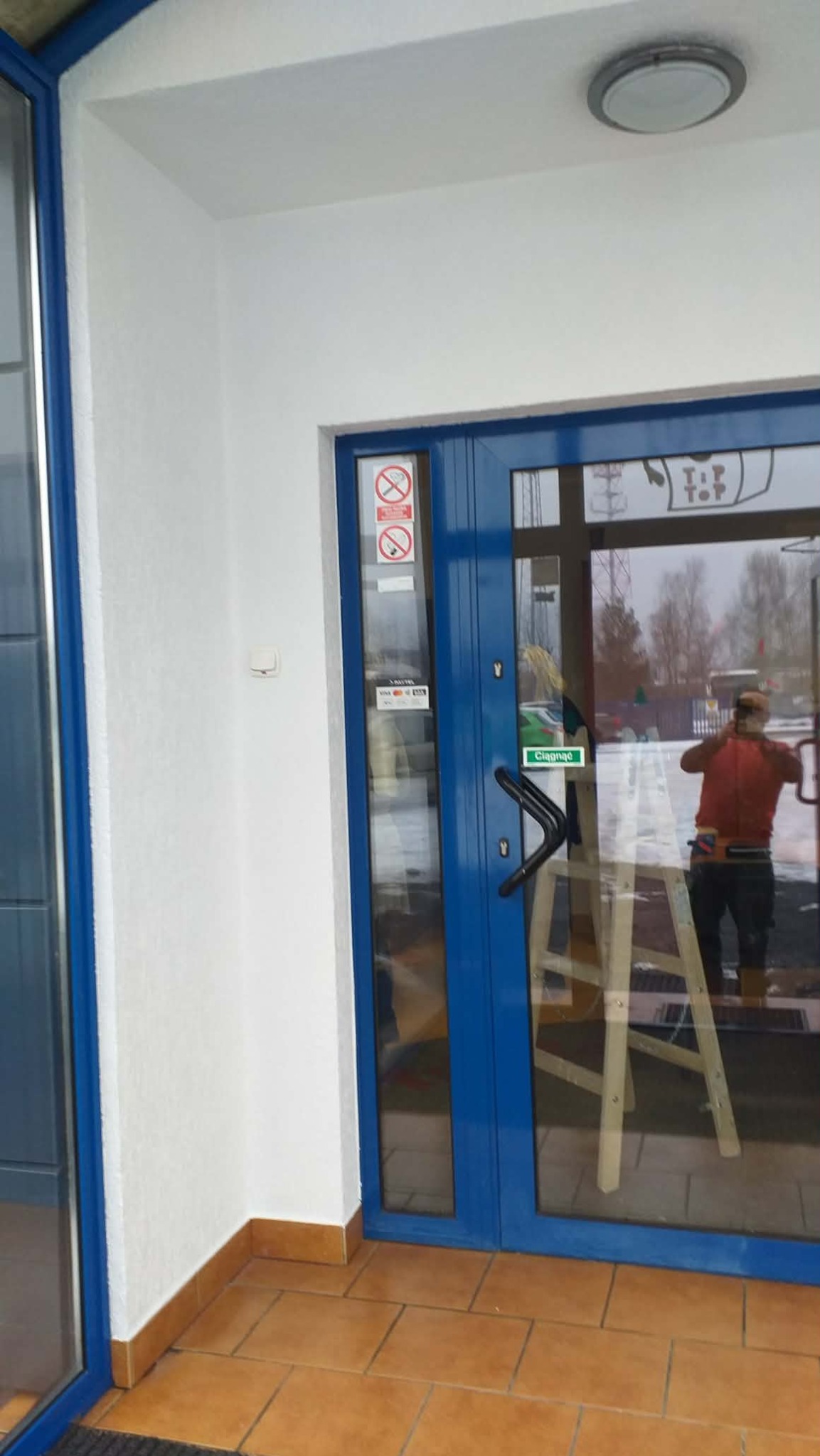 Biała elewacja budynku z niebieskimi drzwiami wejściowymi. W odbiciu szyby widoczny pracownik na drabinie. Podłoga wyłożona brązowymi płytkami.