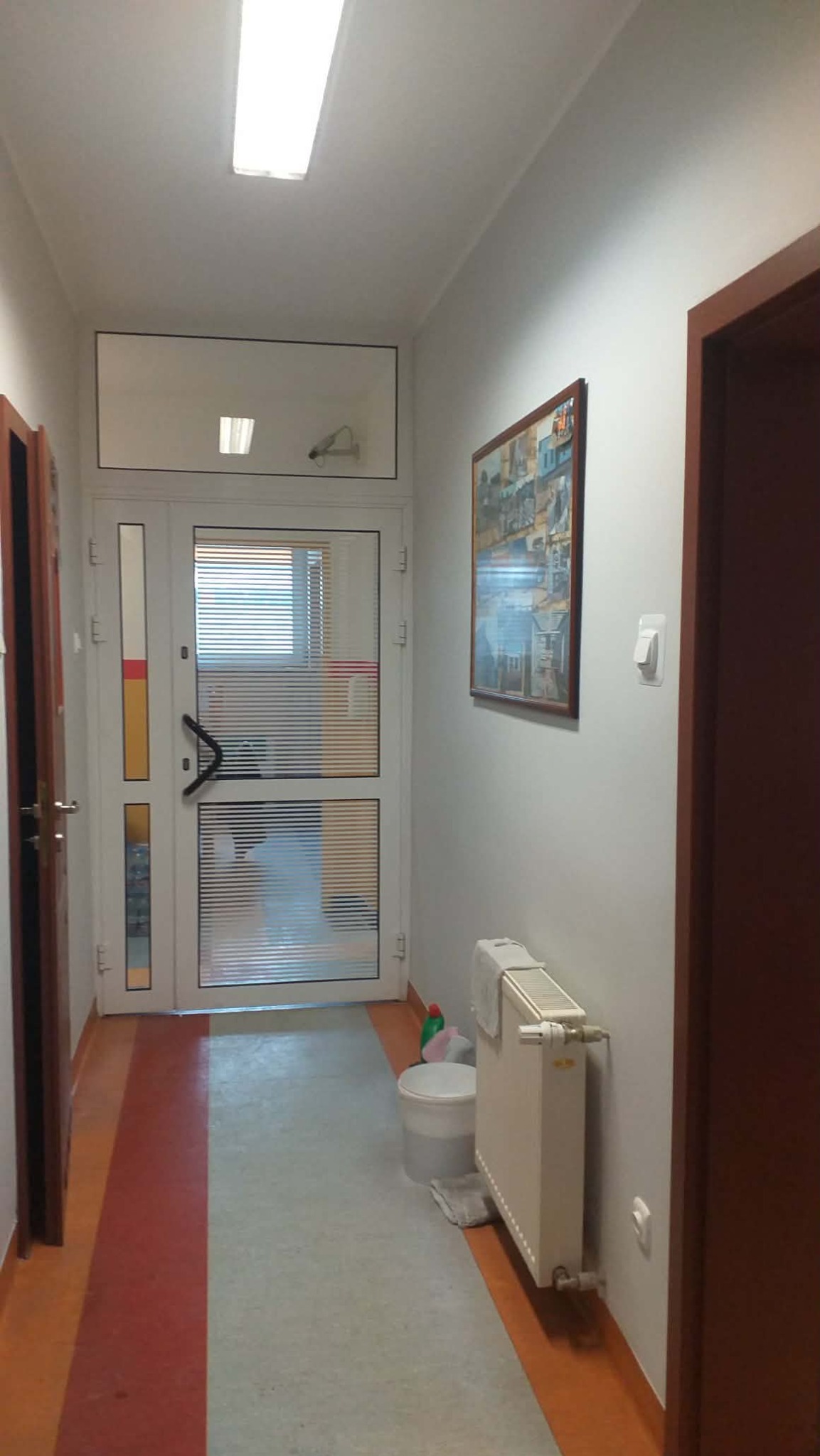Korytarz po odświeżeniu: białe ściany, obraz na ścianie, białe drzwi z żaluzjami, grzejnik. Podłoga z szarym pasem na środku i pomarańczowymi brzegami.