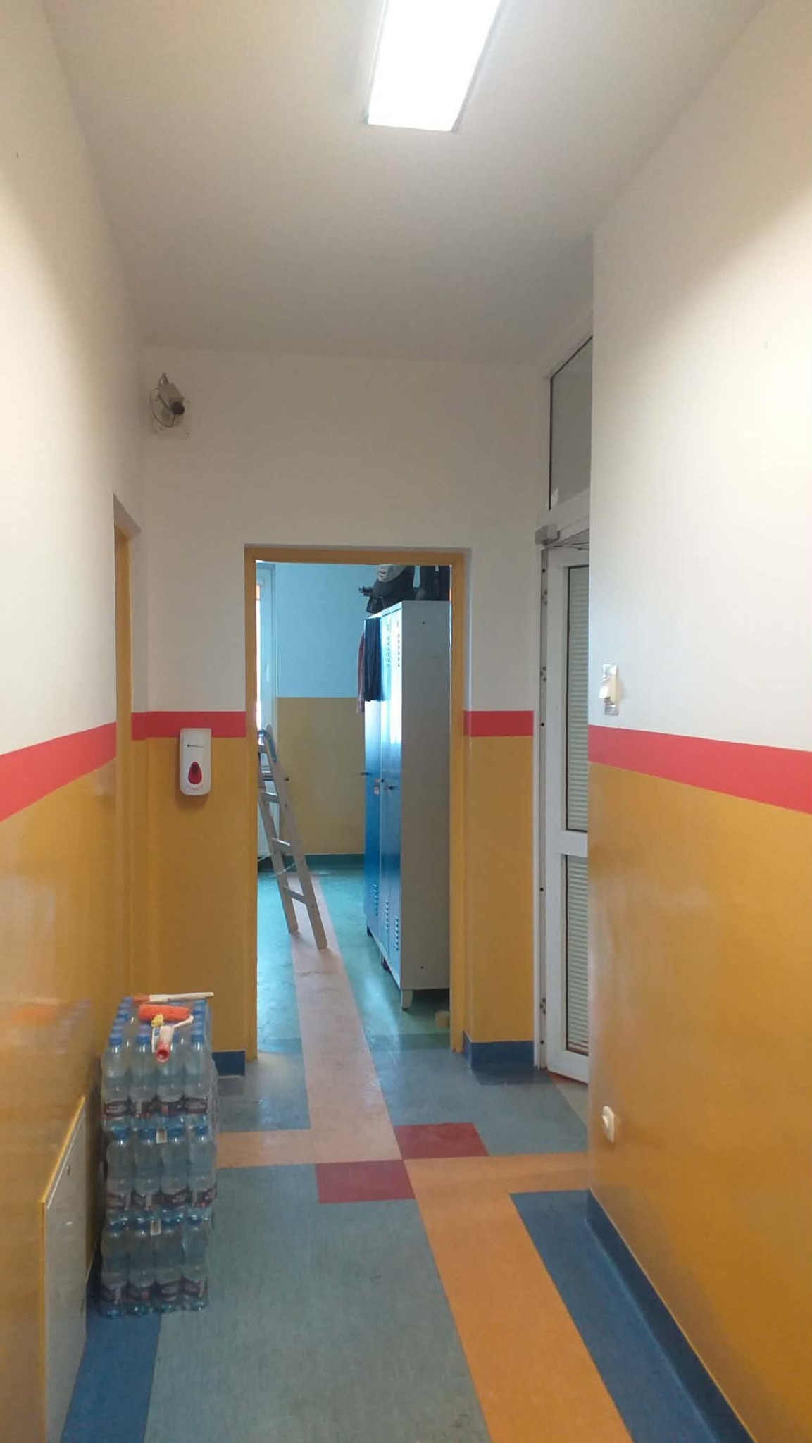 Korytarz po malowaniu ścian na żółto i biało z czerwonym paskiem. Na podłodze wzór w kwadraty. Widoczny dozownik i butelki z wodą.