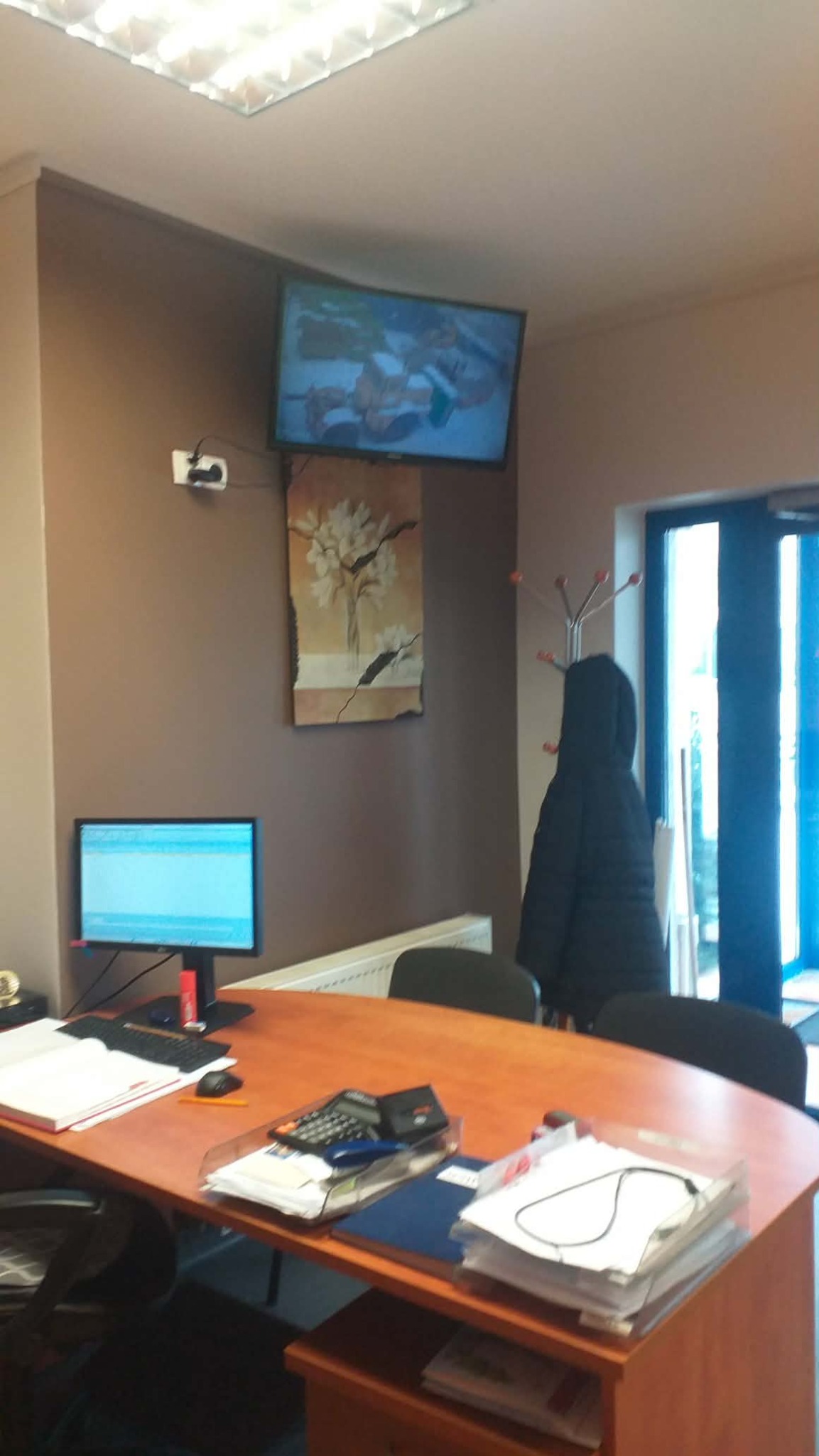 Biuro z brązową ścianą, telewizorem i obrazem. Na biurku komputer, kalkulator i dokumenty. W tle wieszak na ubrania i przeszklone drzwi.