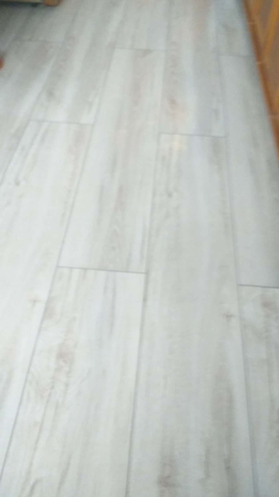 Jasne panele podłogowe ułożone wzdłuż, imitujące drewno, z widoczną strukturą słojów i delikatnymi, szarymi przebarwieniami. Widok z góry, lekko rozmyty.