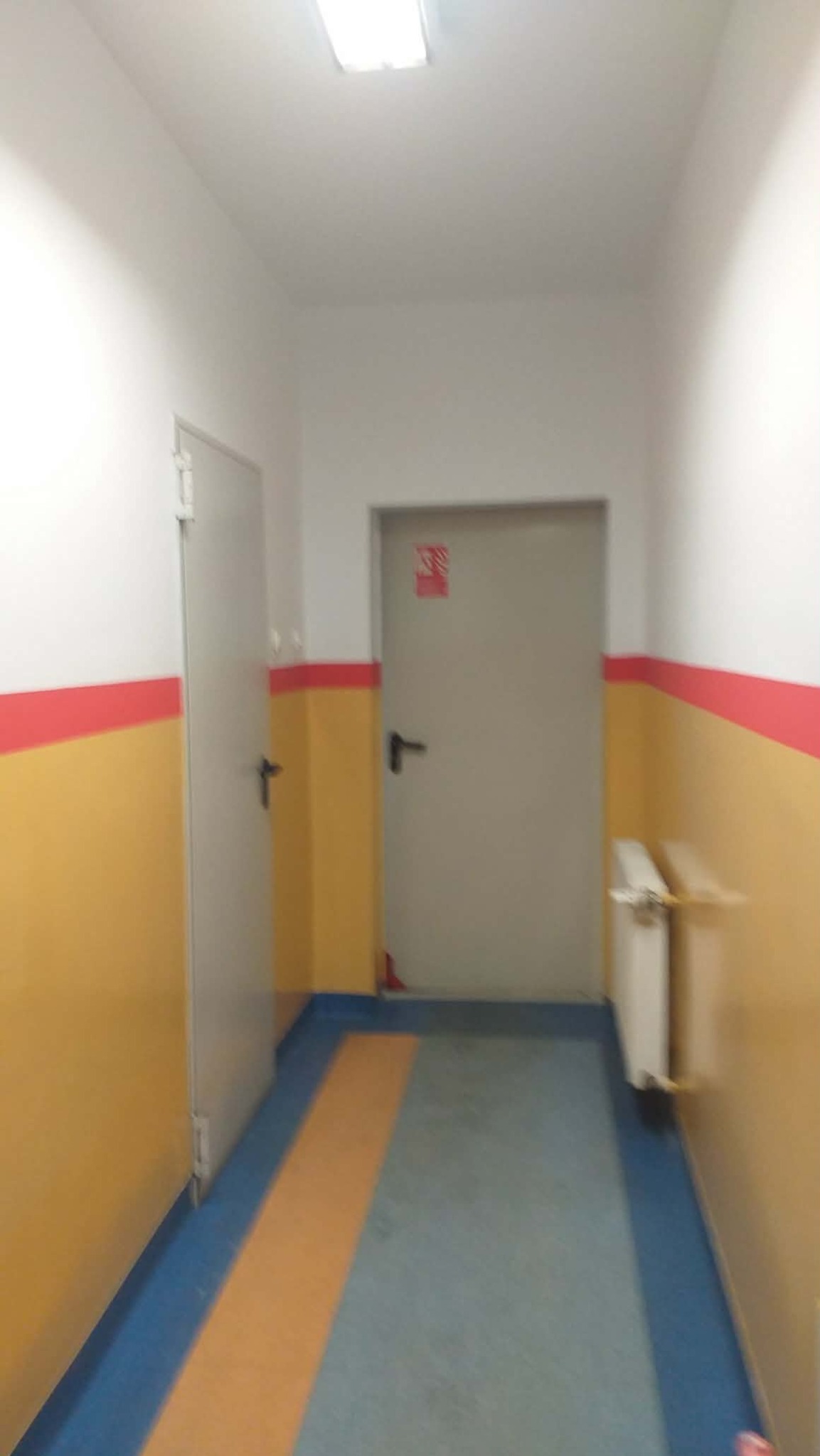 Korytarz po odświeżeniu: żółte ściany z czerwonym pasem, szare drzwi i podłoga w odcieniach niebieskiego i pomarańczowego. Widoczny grzejnik i oświetlenie sufitowe.