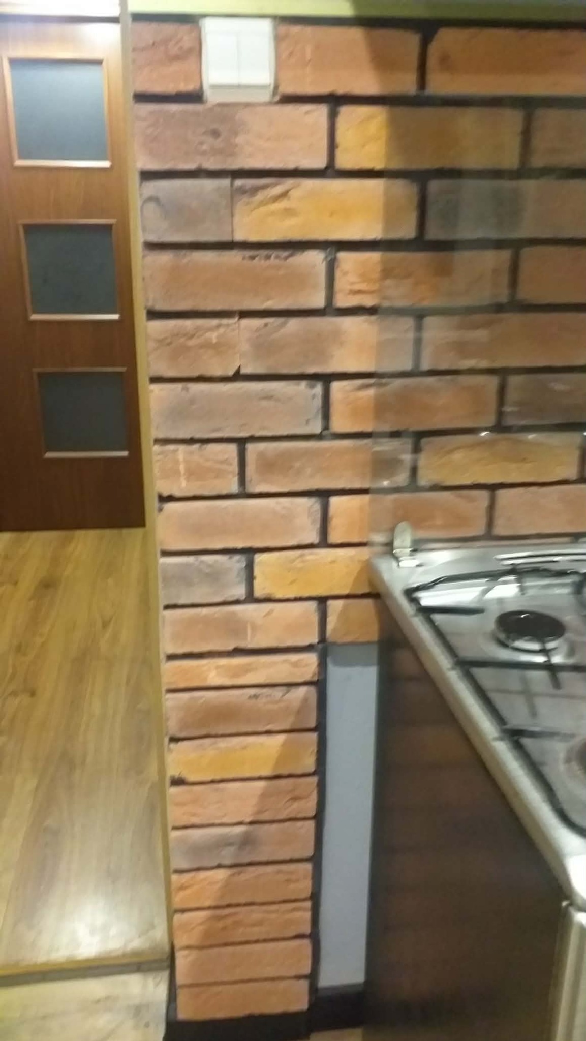 Dekoracyjna ściana z cegły w kuchni, widoczny fragment kuchenki gazowej i drewniane drzwi z przeszkleniami. Ciepłe odcienie brązu i pomarańczy na cegłach.