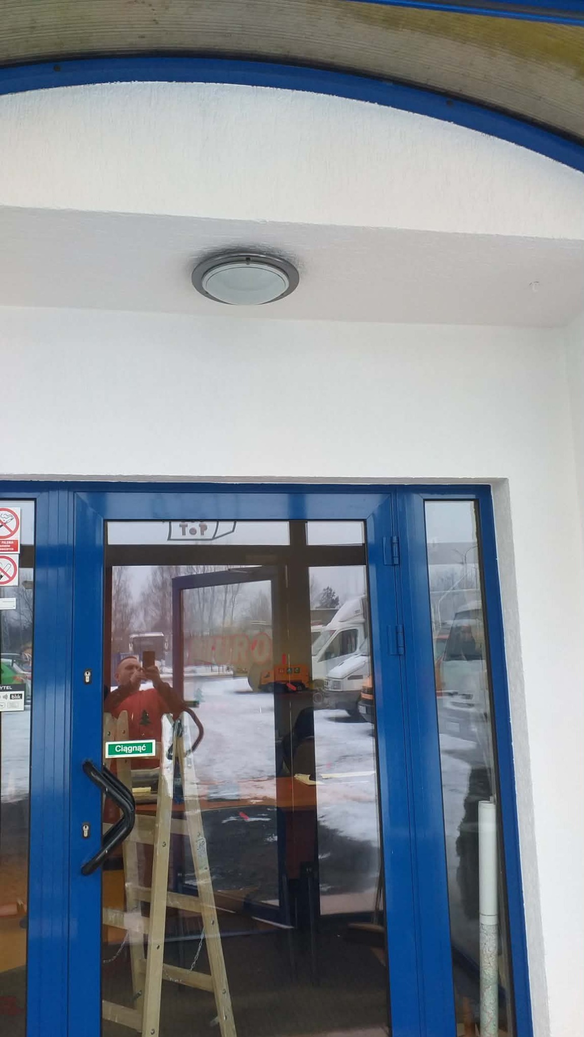Błękitne drzwi wejściowe po malowaniu, widoczna drabina i odbicie osoby robiącej zdjęcie. Świeżo pomalowana elewacja na biało, okrągła lampa sufitowa.
