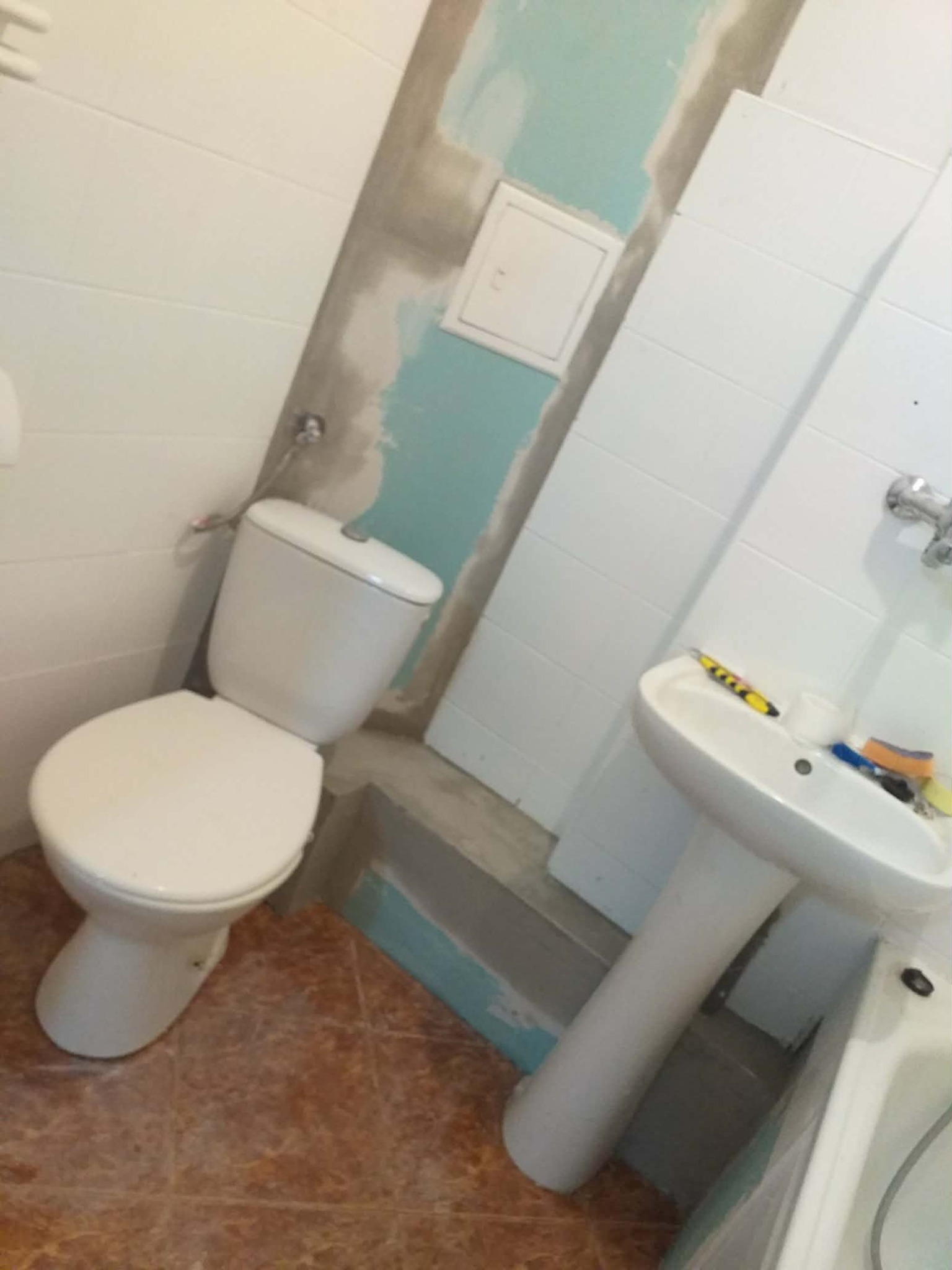 Wnętrze łazienki w trakcie remontu: biała umywalka na postumencie, toaleta i fragment wanny. Widoczne ślady szpachlowania na ścianach i płytki podłogowe w odcieniach brązu.