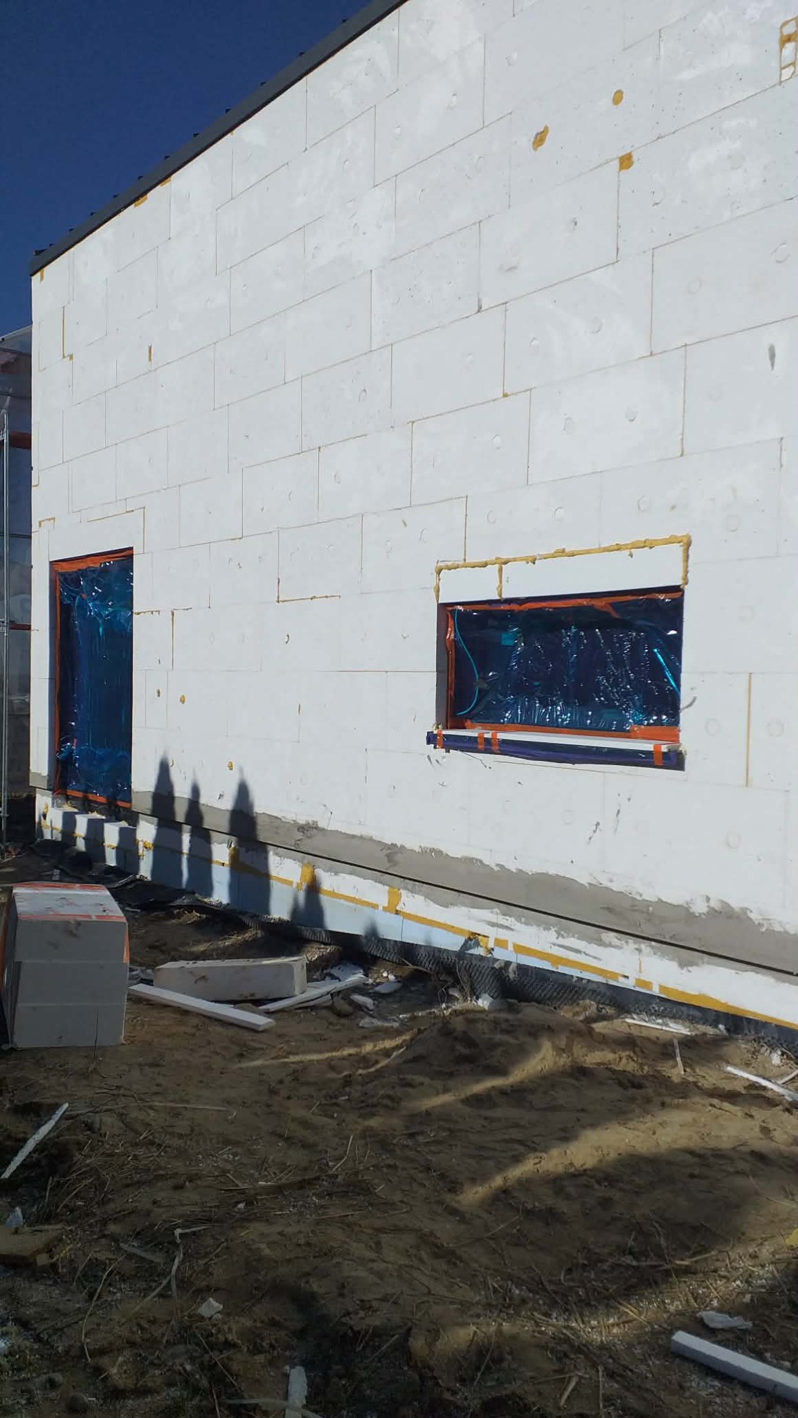 Ściana budynku w trakcie ocieplania, widoczne białe bloki styropianowe, zabezpieczone okna folią, przygotowane podłoże. Etap prac ociepleniowych.