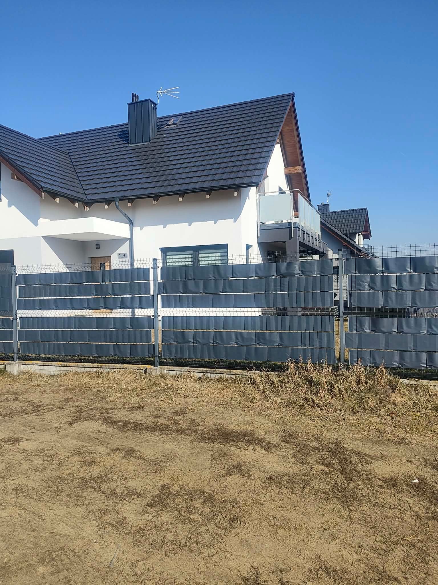 Nowoczesny dom jednorodzinny z szarą elewacją i ciemnym dachem, ogrodzony panelowym płotem z szarymi przesłonami. Błękitne niebo w tle.