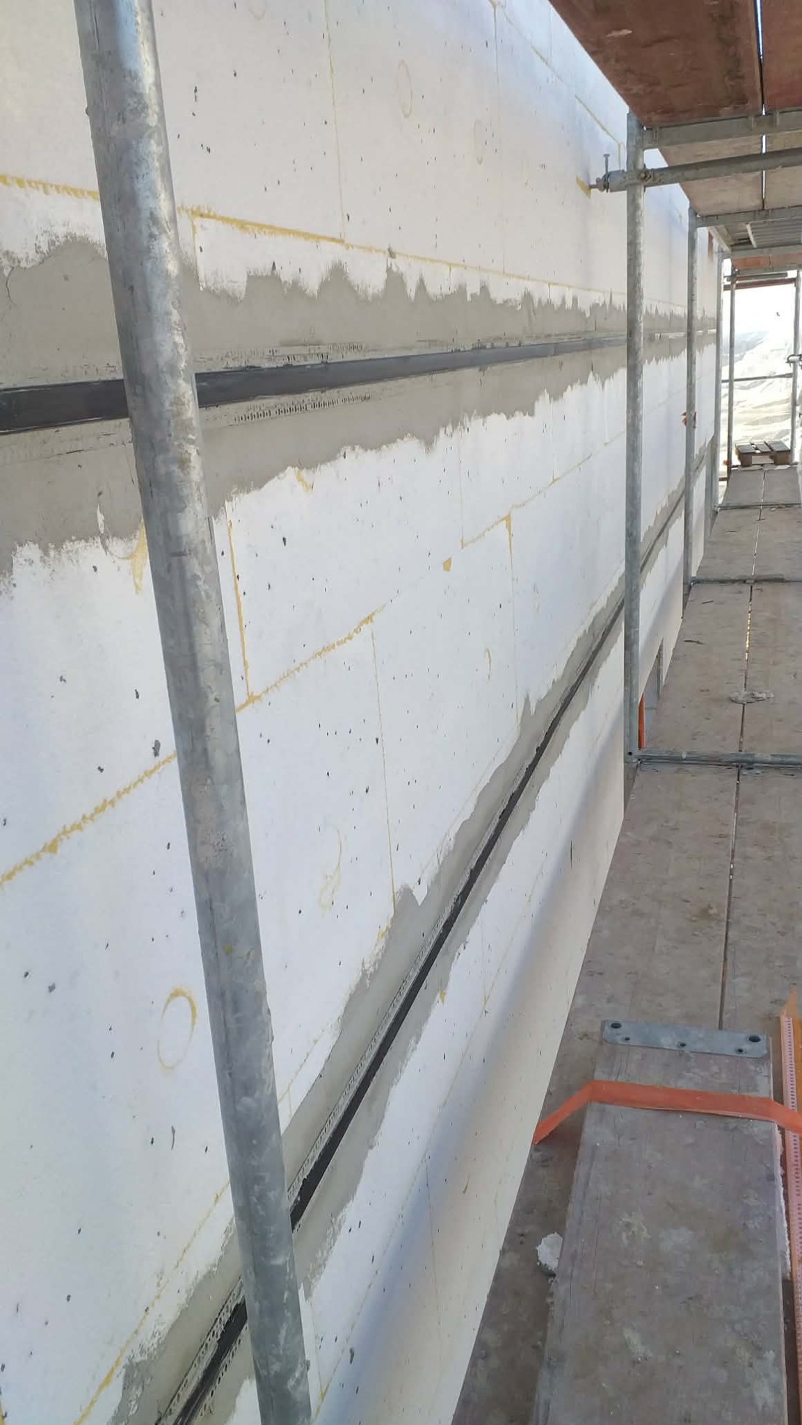 Ocieplona ściana budynku w trakcie prac wykończeniowych, widoczne rusztowania i warstwa izolacyjna z wypełnionymi spoinami. Ujęcie z dołu, perspektywa z boku.