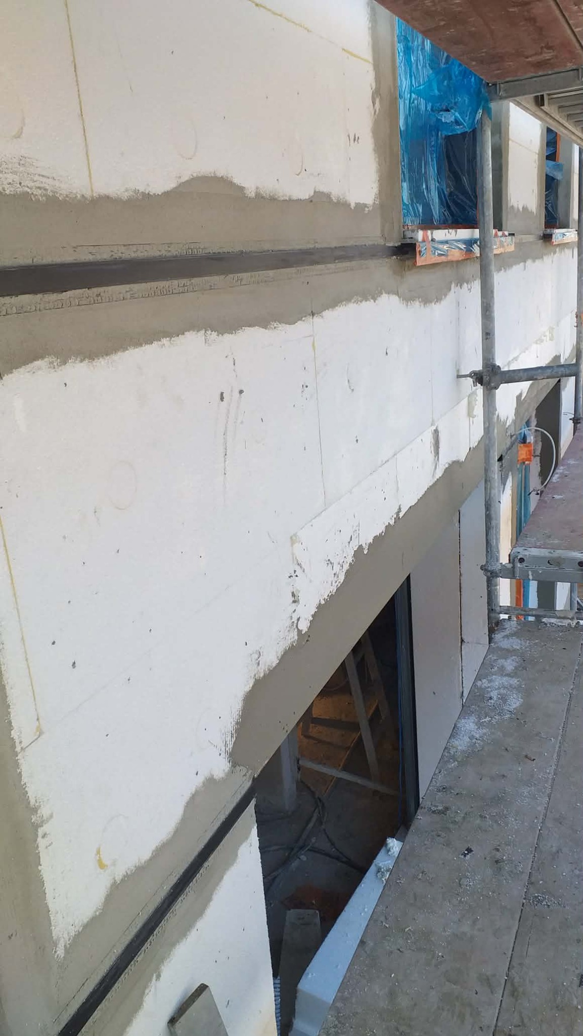 Ocieplona ściana budynku w trakcie prac wykończeniowych, widoczne rusztowania i zabezpieczone okna folią. Szara warstwa szpachli na białym styropianie.
