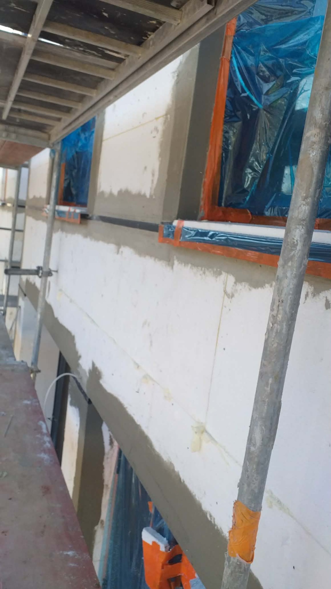 Ocieplanie budynku w trakcie prac. Widoczne rusztowanie, okna zabezpieczone folią, ściana z nałożoną warstwą izolacyjną i szpachlą. Ujęcie z dołu, z bliska.