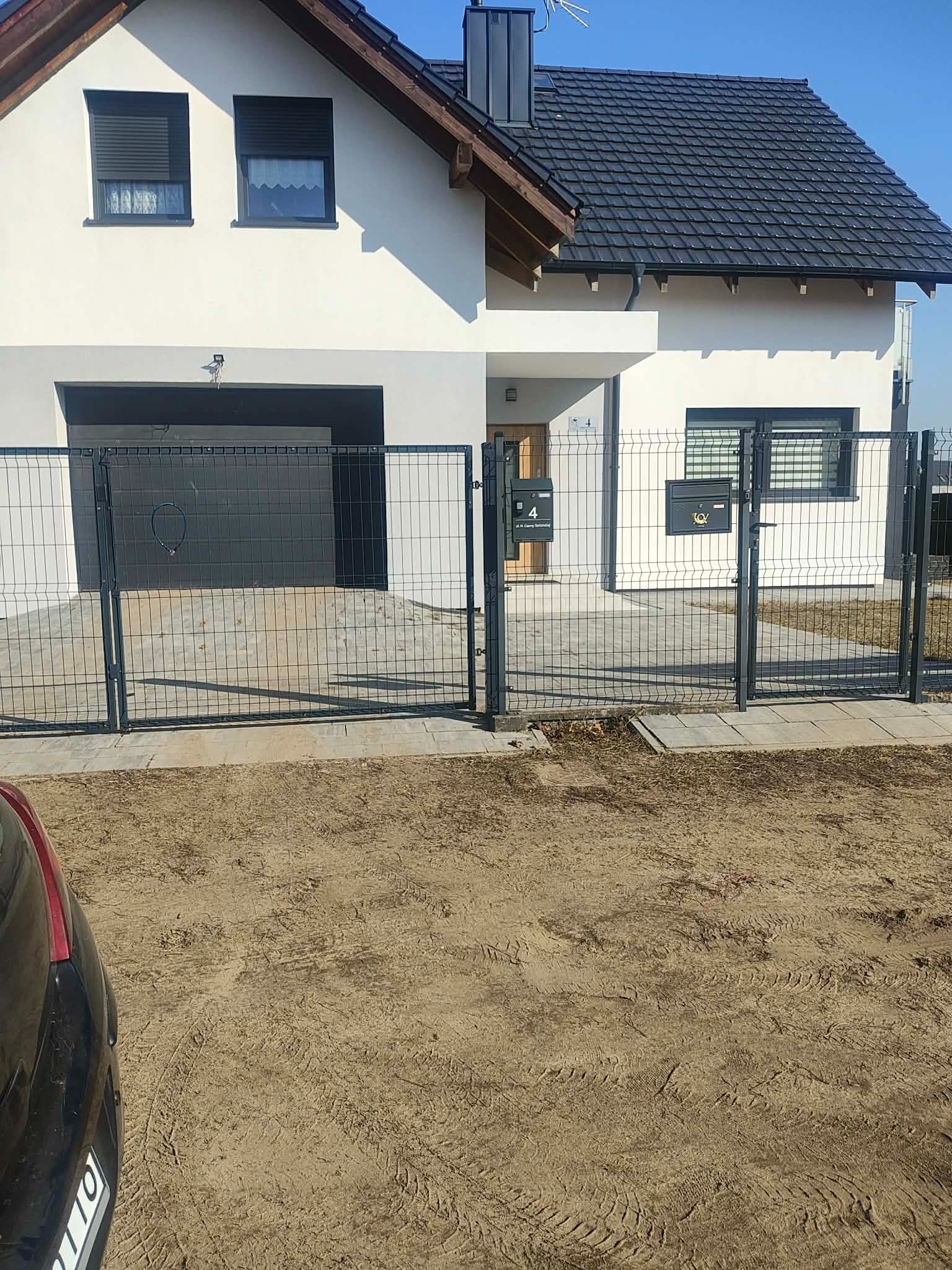 Nowoczesny dom jednorodzinny z szarą bramą garażową i metalowym ogrodzeniem panelowym. Widoczne skrzynki pocztowe przy furtce. Jasna elewacja i ciemna dachówka.