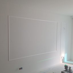 Remonty wykonczenia Krzysztof Barciszewski - Białe ściany z dekoracyjnymi listwami w prostokątnych ramach, wyznaczane laserem. Widoczny fragment osoby i narzędzia. Wnętrze w trakcie wykańczania.