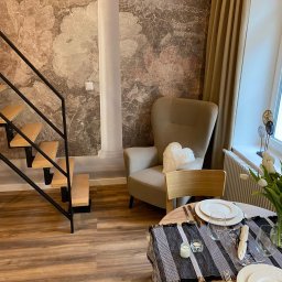 Remonty wykonczenia Krzysztof Barciszewski - Stylowe wnętrze: drewniane schody z czarną balustradą, fotel z poduszką w kształcie serca, okrągły stół z nakryciem i bukietem białych tulipanów na tle ściany z motywem kwiatowym.
