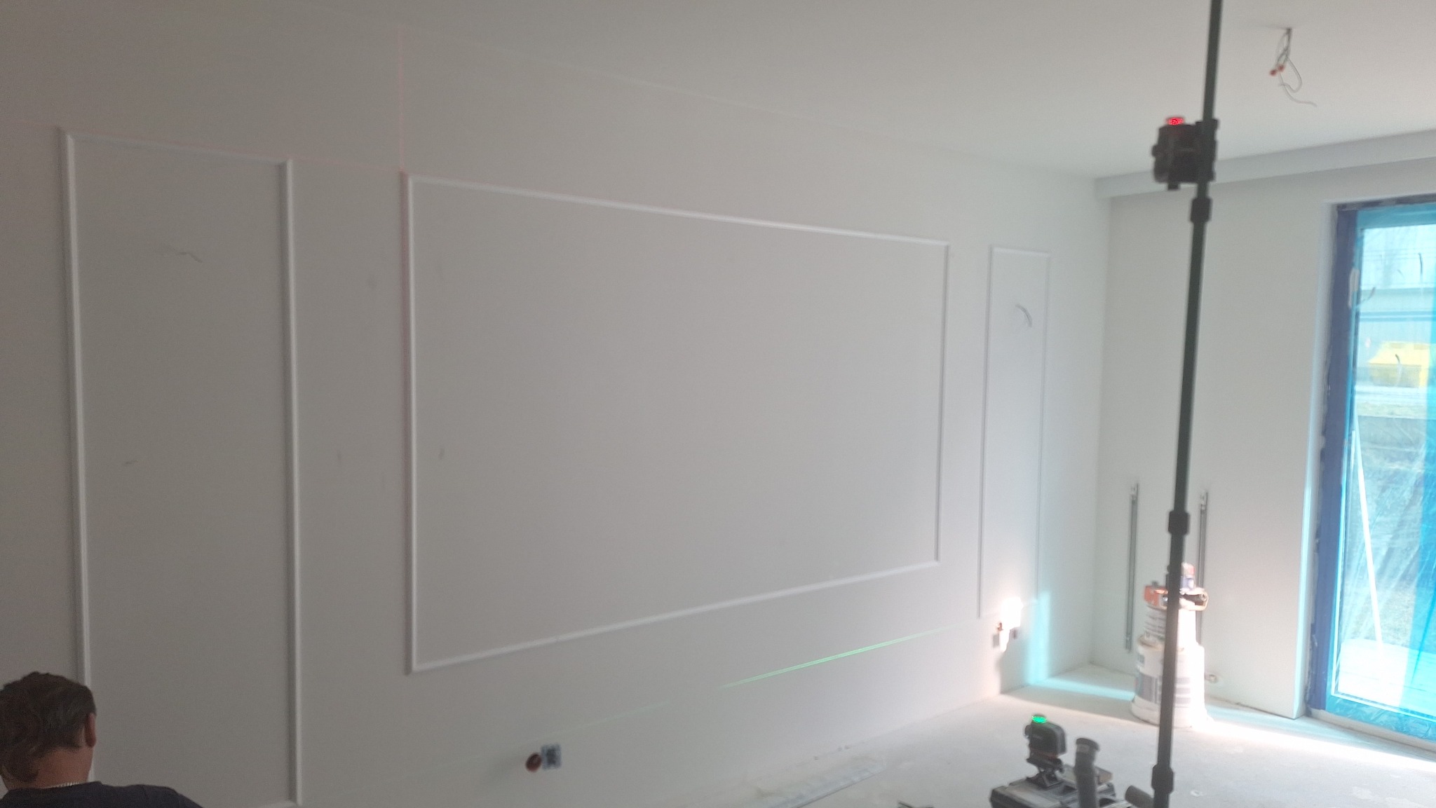 Białe ściany z dekoracyjnymi listwami w prostokątnych ramach, wyznaczane laserem. Widoczny fragment osoby i narzędzia. Wnętrze w trakcie wykańczania.