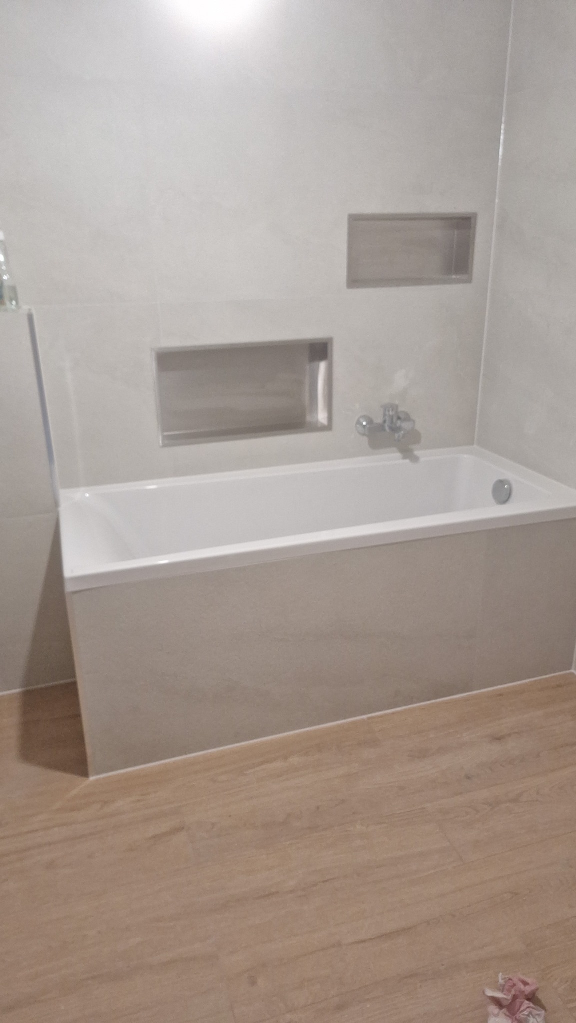 Wykończona łazienka z wanną w zabudowie, jasnymi płytkami i drewnopodobną podłogą. W ścianie wnęki wykończone płytkami. Minimalistyczny design.