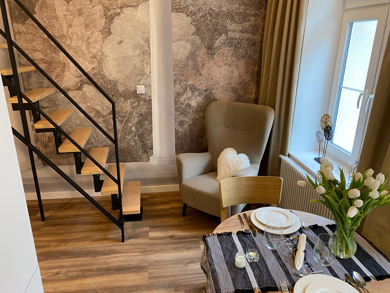 Stylowe wnętrze: drewniane schody z czarną balustradą, fotel z poduszką w kształcie serca, okrągły stół z nakryciem i bukietem białych tulipanów na tle ściany z motywem kwiatowym.
