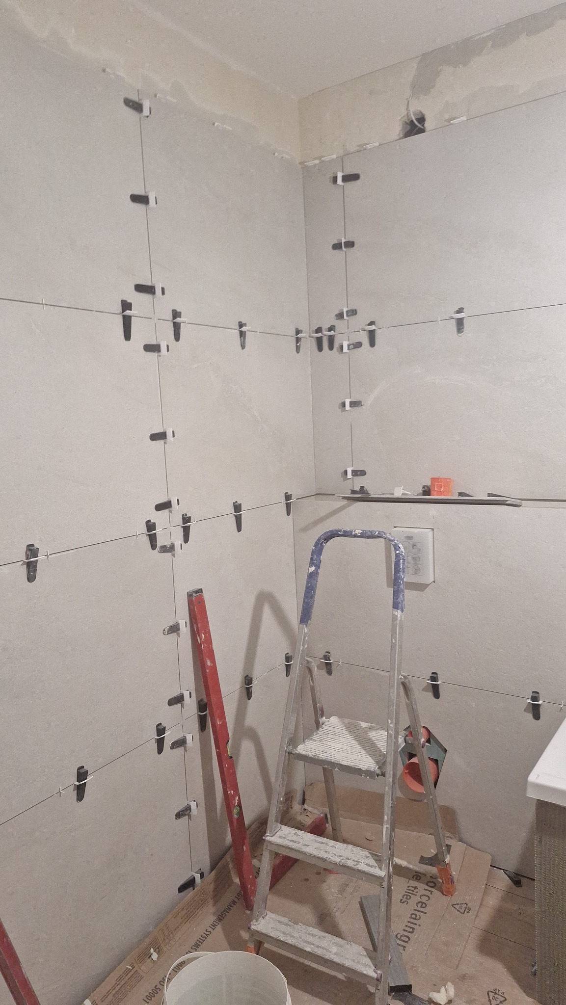 Ściana w trakcie układania płytek, z użyciem systemu poziomowania. Widoczna drabina, poziomica i wiadro. Jasne płytki, szare fugi, etap prac wykończeniowych.