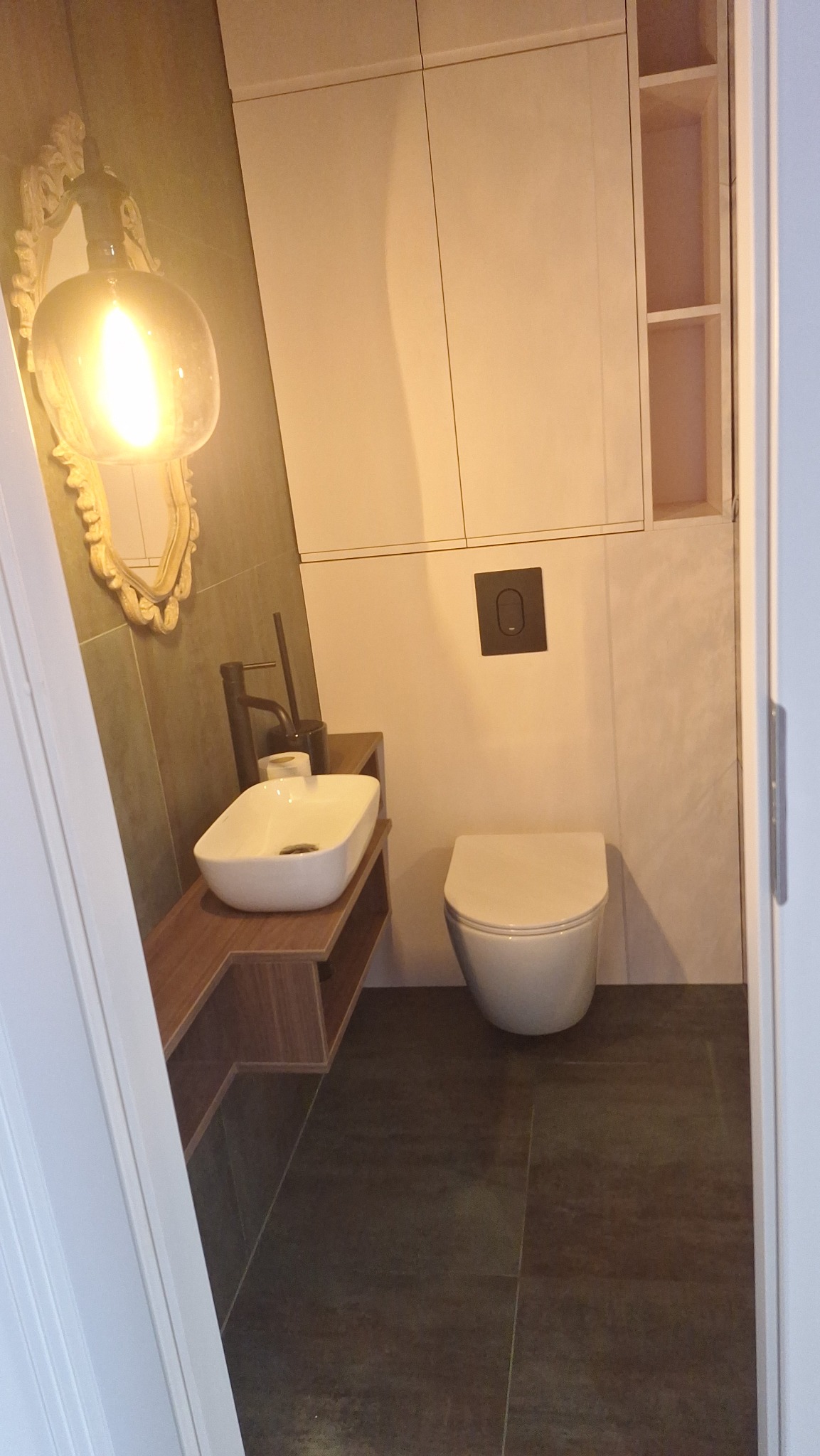 Nowoczesna łazienka z umywalką na drewnianym blacie, ozdobnym lustrem i podwieszaną toaletą. Minimalistyczny design, szare płytki podłogowe, białe szafki wiszące.