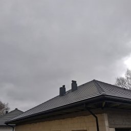 Usługi budowlane KLM - Nowy, grafitowy dach z ceramicznej dachówki karpiówki, z czarnymi rynnami i kominami, na tle pochmurnego nieba. Widok z dołu, fragment ściany budynku w surowym stanie.