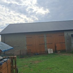 Usługi budowlane KLM - Szary dach z blachy trapezowej na budynku gospodarczym w Zduńskiej Woli. Widoczne drewniane wrota i fragment podwórka z zielenią. Pochmurne niebo w tle.
