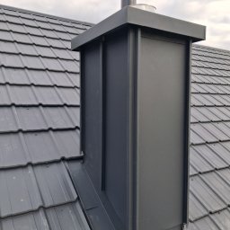 Usługi budowlane KLM - Szary, metalowy komin na dachu z blachy trapezowej. Komin z czarnym daszkiem wentylacyjnym. Widok z dołu, pochmurne niebo w tle. Obróbki blacharskie.