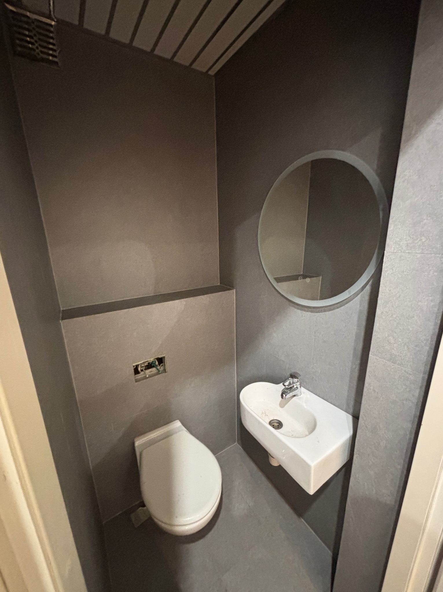 Szara toaleta po remoncie: umywalka, okrągłe lustro z podświetleniem, wisząca muszla klozetowa. Minimalistyczny design, płytki imitujące kamień na ścianach i podłodze.