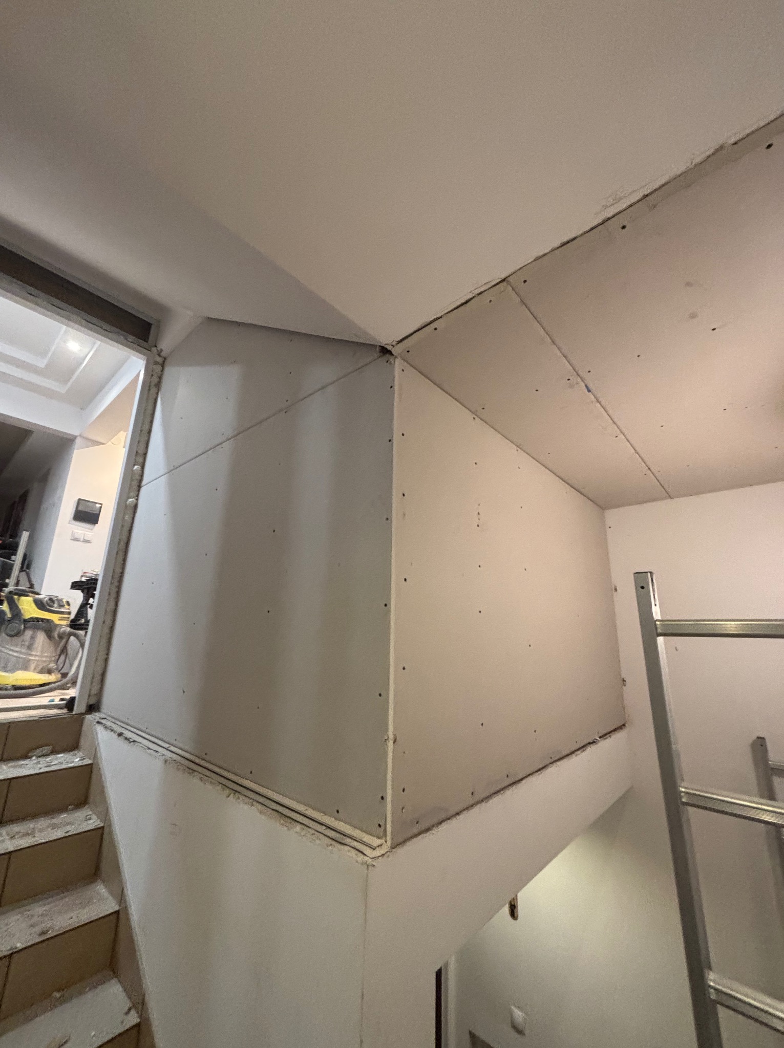 Montaż płyt karton-gips na ścianie przy schodach, widoczne łączenia płyt i drabina. W tle odkurzacz przemysłowy i fragment wnętrza mieszkania.