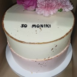 Klaudia Stefańska - Dwupoziomowy tort urodzinowy z kremem, złotymi akcentami i kwiatami, z napisem '50 Moniki', umieszczony w kartonowym pudełku.