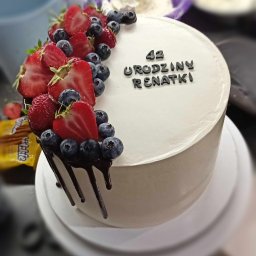 Klaudia Stefańska - Tort urodzinowy z kremem, dekorowany świeżymi truskawkami, borówkami i polewą czekoladową. Napis '42 URODZINY RENATKI' wykonany czarną czcionką.