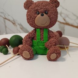 Klaudia Stefańska - Ręcznie wykonany, brązowy miś z modeliny w zielonych ogrodniczkach, otoczony kolorowymi kulkami i patyczkami na białym tle. Dekoracja tortu.