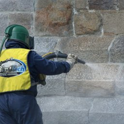 BLAST TECH SPÓŁKA Z OGRANICZONĄ ODPOWIEDZIALNOŚCIĄ - Pracownik w hełmie i kamizelce z logo BlastTech czyści kamienną elewację strumieniem wody pod ciśnieniem, usuwając zabrudzenia i odsłaniając czystą powierzchnię.