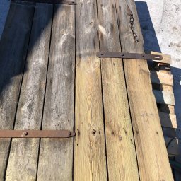 BLAST TECH SPÓŁKA Z OGRANICZONĄ ODPOWIEDZIALNOŚCIĄ - Stare, drewniane wrota z metalowymi zawiasami, częściowo oczyszczone, leżące na palecie. Widoczna różnica w kolorze drewna przed i po czyszczeniu.