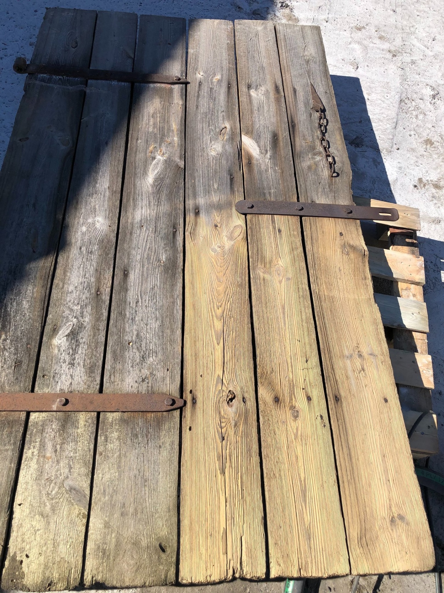 Stare, drewniane wrota z metalowymi zawiasami, częściowo oczyszczone, leżące na palecie. Widoczna różnica w kolorze drewna przed i po czyszczeniu.