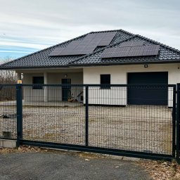 Polstal - Nowoczesny dom jednorodzinny z panelami słonecznymi na dachu, widziany zza metalowej bramy panelowej w kolorze grafitowym.