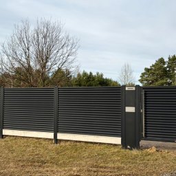 Polstal - Nowoczesne, poziome panele ogrodzeniowe w kolorze antracytowym, osadzone na betonowej podmurówce. Widoczna brama z wbudowaną skrzynką na listy. Tło stanowią drzewa.