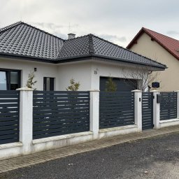 Polstal - Nowoczesne ogrodzenie panelowe w kolorze grafitowym z białymi słupkami, otaczające posesję z domem jednorodzinnym. Widoczna brama wjazdowa i furtka.