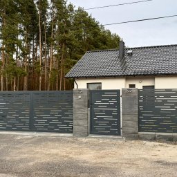 Polstal - Nowoczesna, szara brama wjazdowa i przęsła ogrodzeniowe z poziomymi, ażurowymi wycięciami, osadzone na betonowych słupkach. W tle dom i las.