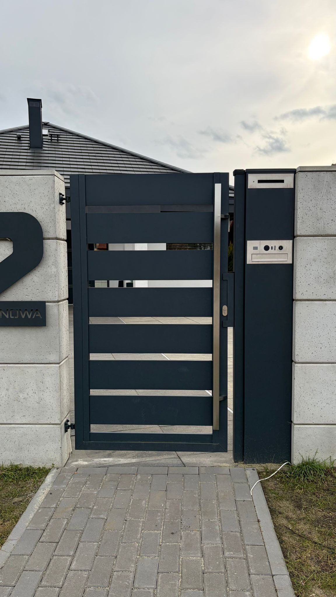 Nowoczesna, antracytowa brama posesyjna z poziomymi szczeblami, wkomponowana w betonowe słupki z numerem domu i skrzynką pocztową. Brukowana ścieżka prowadzi do wejścia.