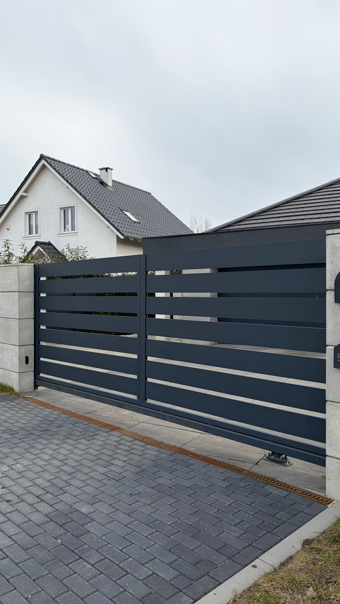 Nowoczesna, przesuwna brama w kolorze antracytu z poziomymi panelami, zamontowana na posesji z kostką brukową i domem w tle. Minimalistyczny design.