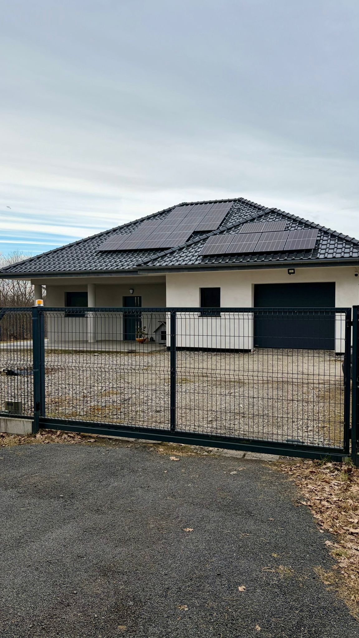 Nowoczesny dom jednorodzinny z panelami słonecznymi na dachu, widziany zza metalowej bramy panelowej w kolorze grafitowym.