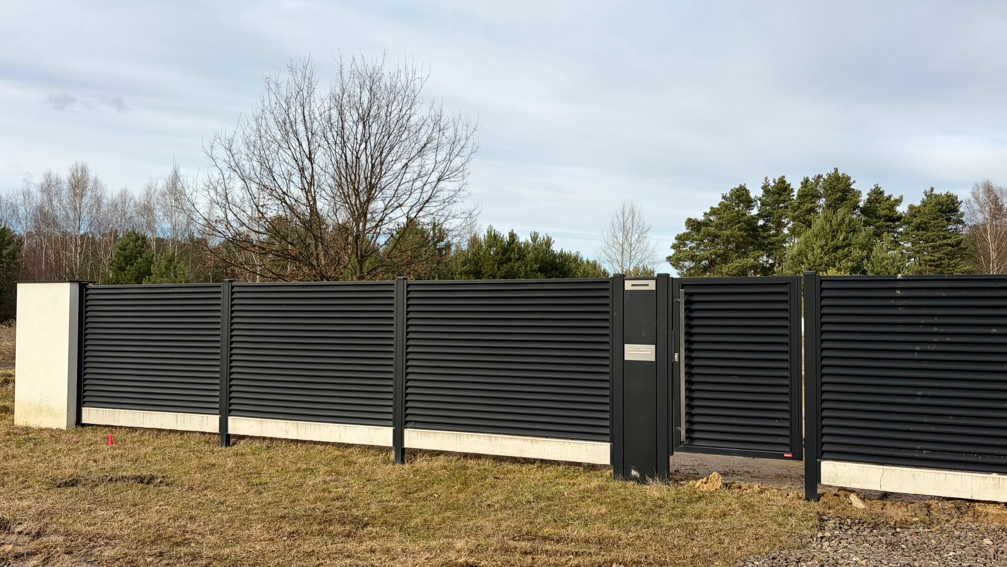 Nowoczesne, poziome panele ogrodzeniowe w kolorze antracytowym, osadzone na betonowej podmurówce. Widoczna brama z wbudowaną skrzynką na listy. Tło stanowią drzewa.